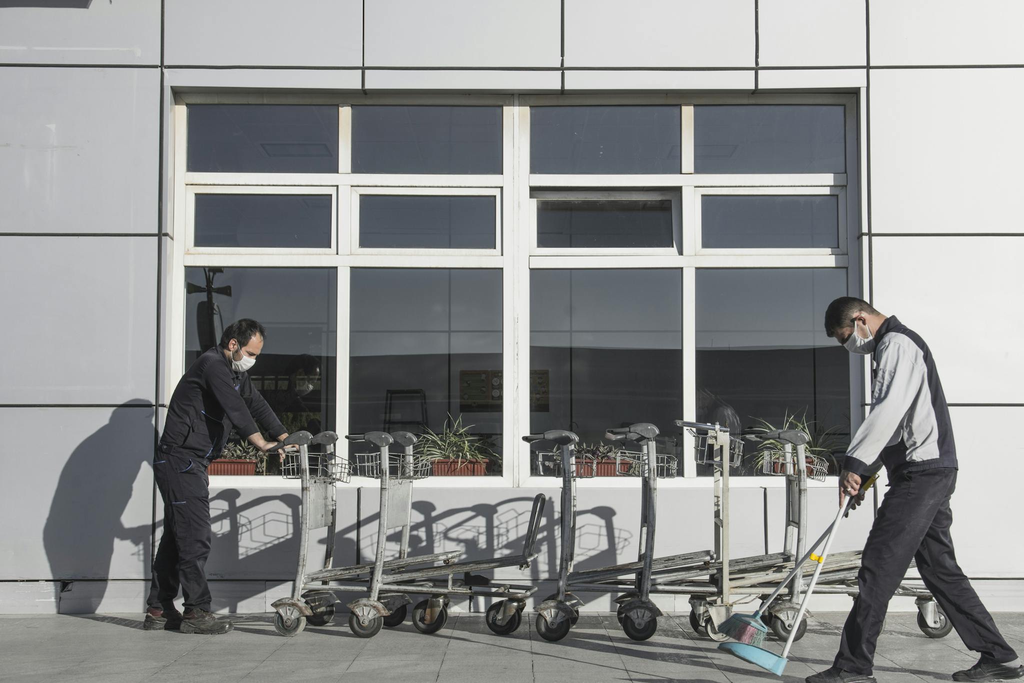 Two masked workers clean and organize trolleys outside a modern building, highlighting industrial maintenance.